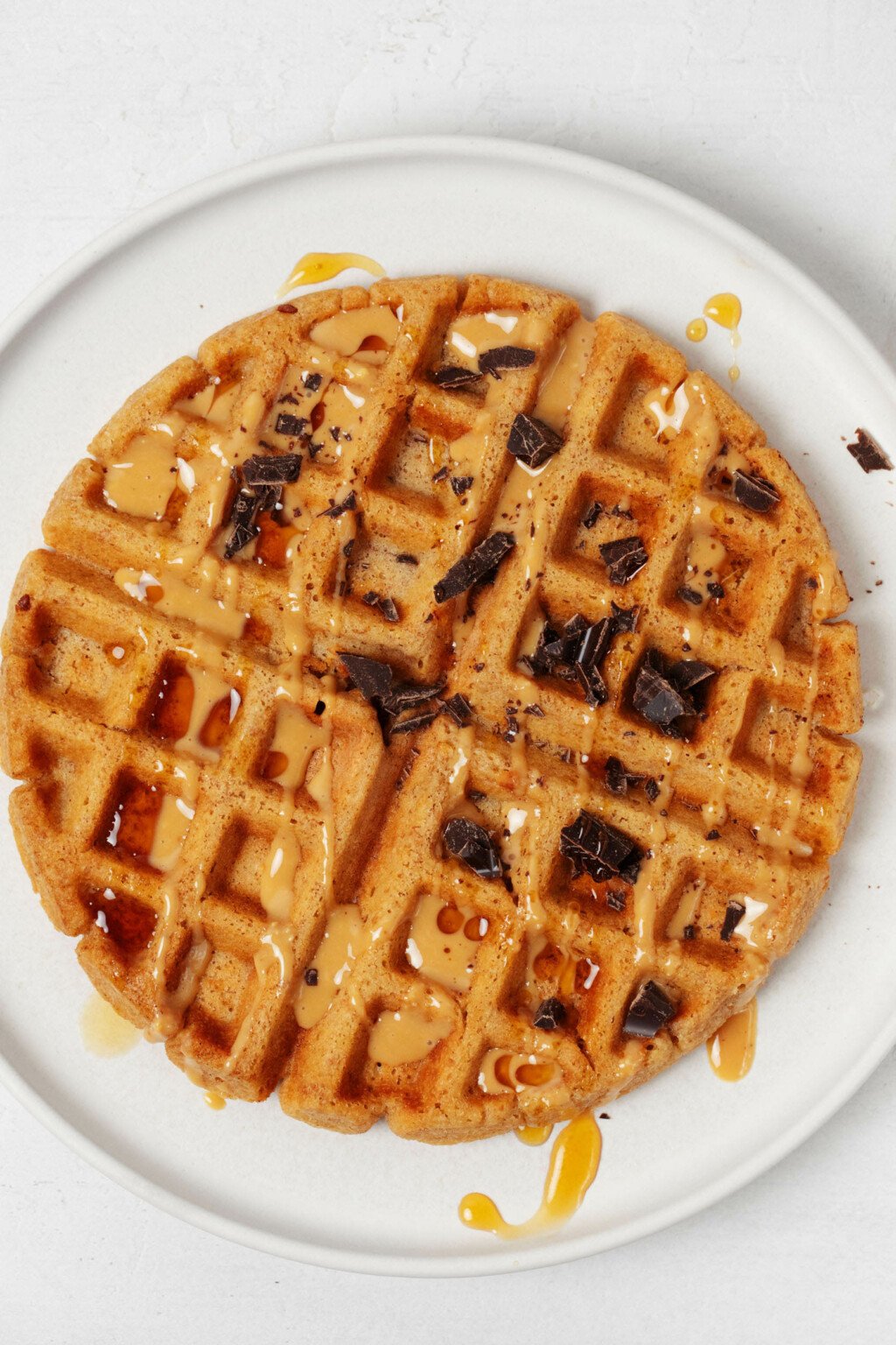 An overhead image of a round, white plate, which is holding a light and crispy vegan peanut butter waffle. The waffle is topped with chopped dark chocolate and a drizzle of peanut butter.