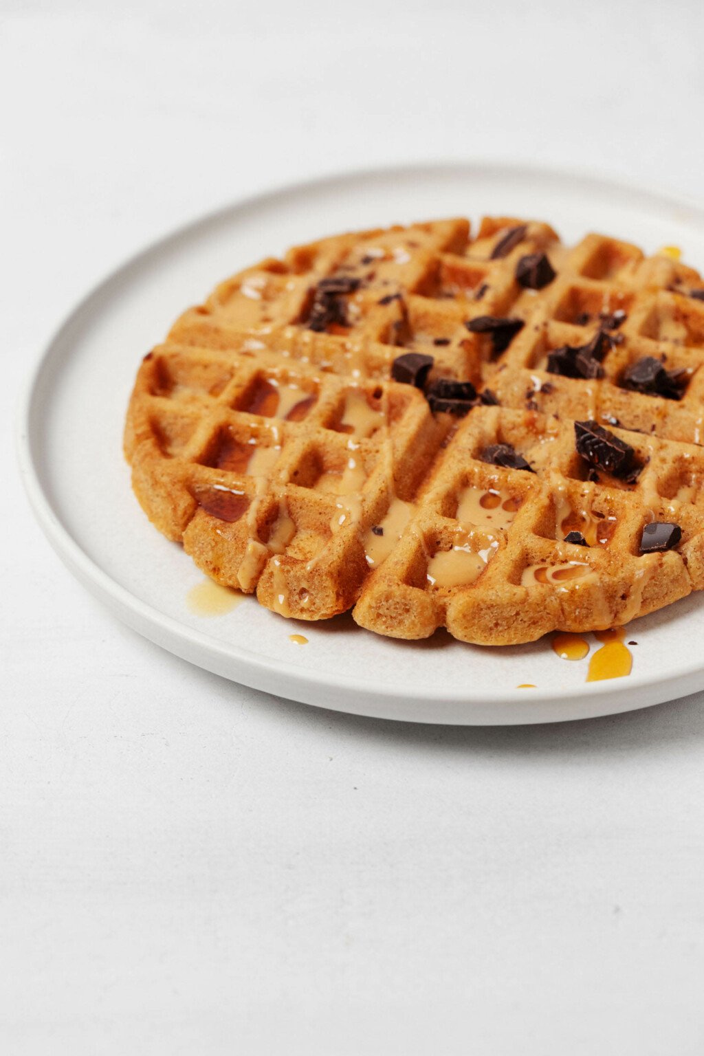 An angled image of a round, white plate, resting on a white surface. The plate holds a Belgian-style vegan peanut butter waffle, which is topped with chocolate and peanut butter drizzle.