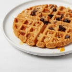 An angled image of a round, white plate, resting on a white surface. The plate holds a Belgian-style vegan peanut butter waffle, which is topped with chocolate and peanut butter drizzle.