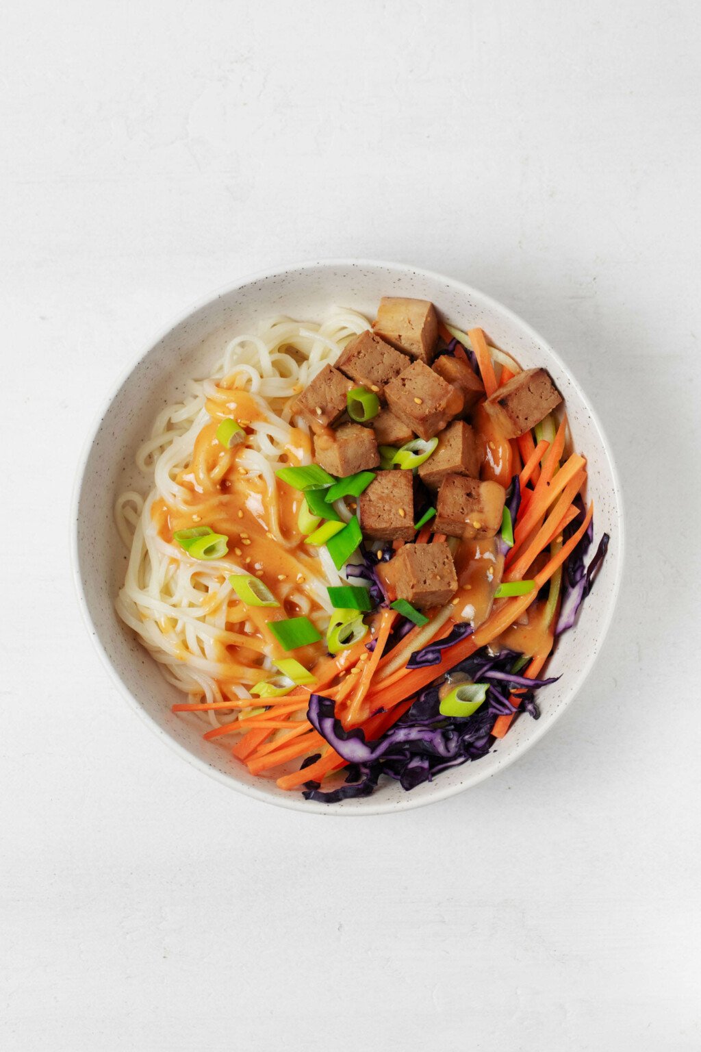 An overhead image of a white bowl filled with cooked noodles, raw carrot and red cabbage, tofu, and a sauce.