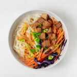 An overhead image of a white bowl filled with cooked noodles, raw carrot and red cabbage, tofu, and a sauce.