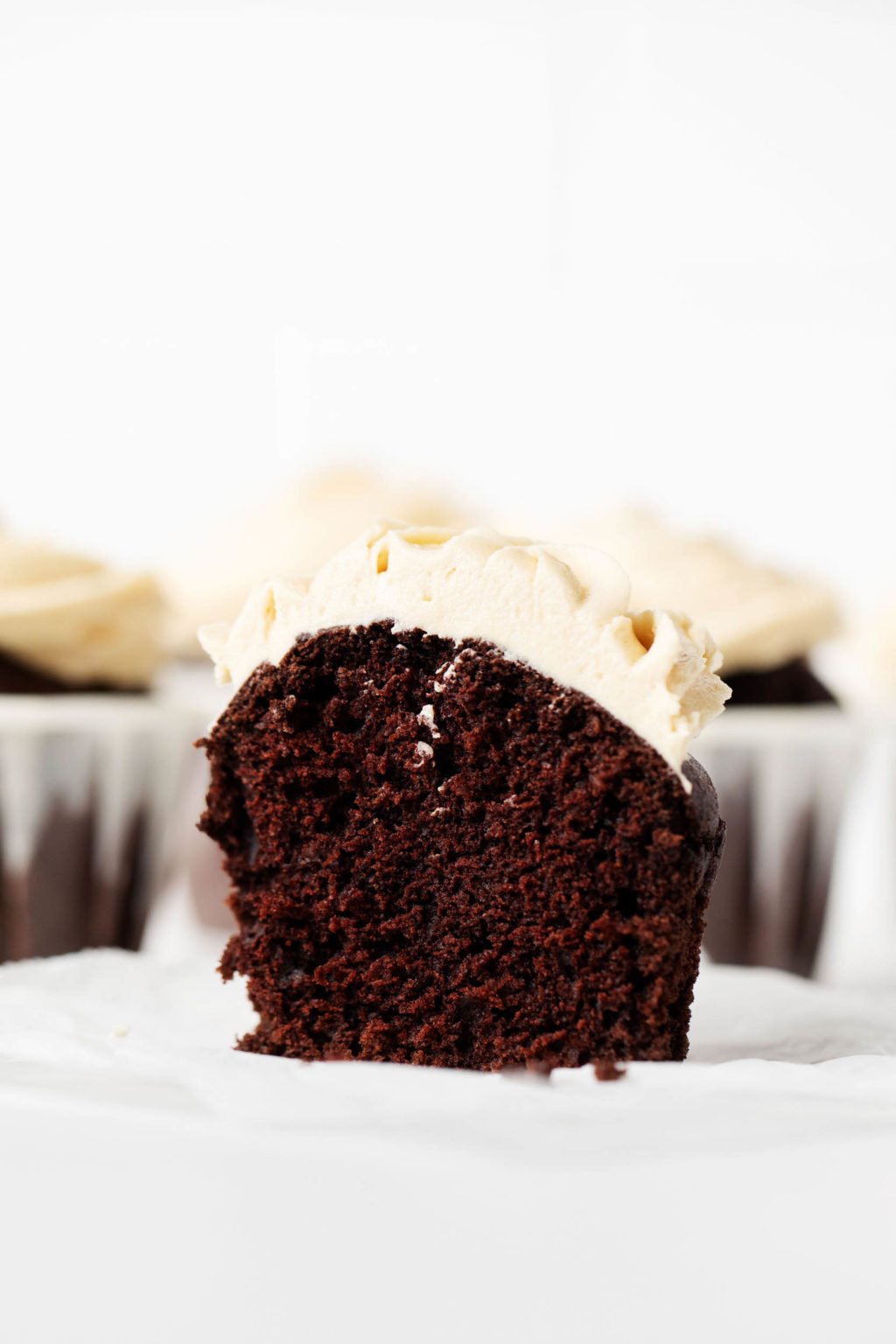 A vegan chocolate peanut butter cupcake has been sliced neatly in half. A cross section of the muffin is visible, with muffins that are still in their liners in the background.