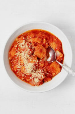 A white bowl is being used to hold a cozy vegan tomato bread soup, topped with plant-based parmesan cheese.