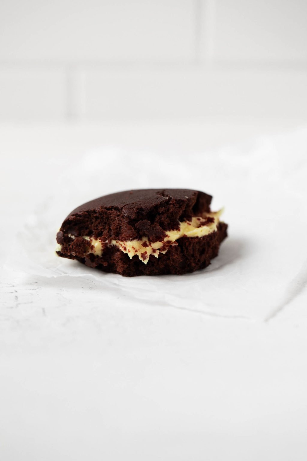 A vegan whoopie pie, made with chocolate cookies and pumpkin spice buttercream filling, just bitten into. The cookie rests on a sheet of parchment.