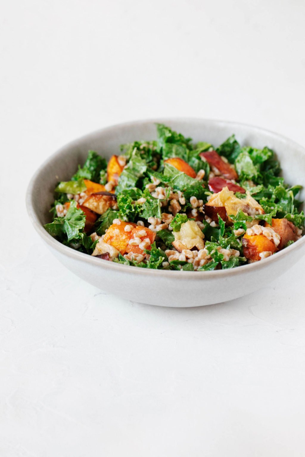 A small, white bowl is filled with a kale, sweet potato, and apple salad. The salad contains grains of farro.