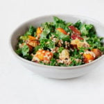 A small, white bowl is filled with a kale, sweet potato, and apple salad. The salad contains grains of farro.