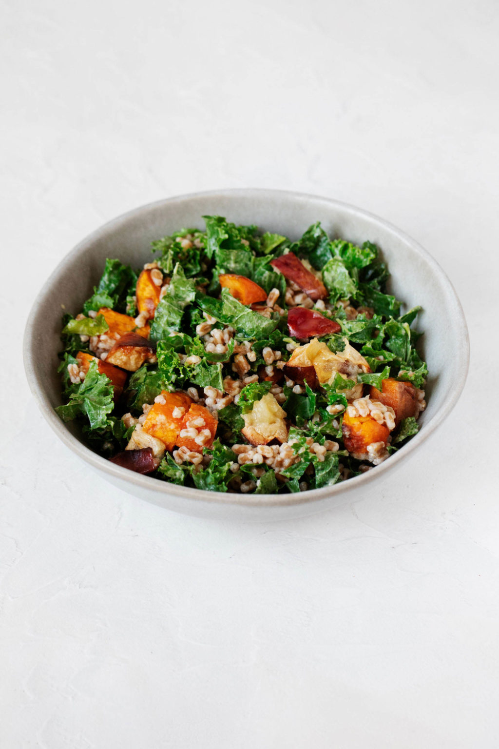 A white, ceramic salad bowl has been filled with a kale and whole grain salad. The salad contains orange sweet potato pieces and has a creamy dressing.