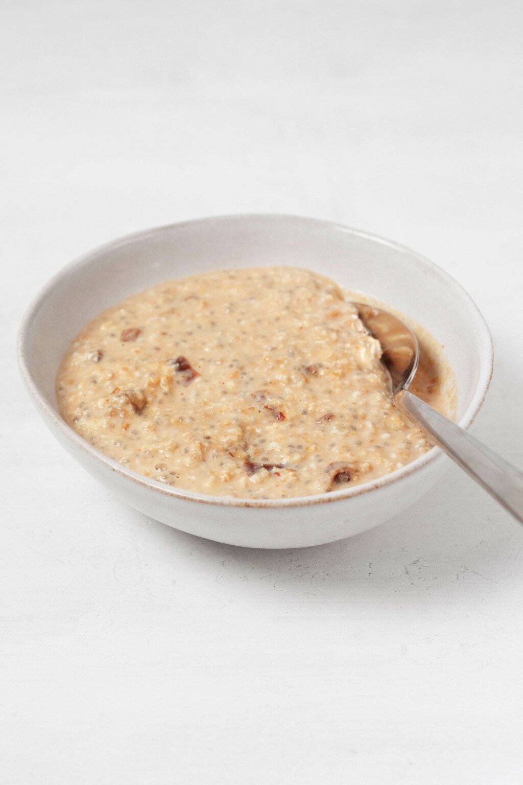 An angled photograph of a bowl of vegan pumpkin overnight oats. A silver teaspoon is resting in the oats, as if they're ready to be eaten.