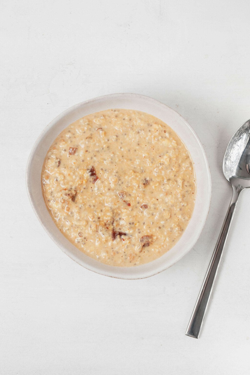 An asymmetrical, white bowl is filled with oats, non-dairy milk, and chia seeds. A teaspoon rests next to it.