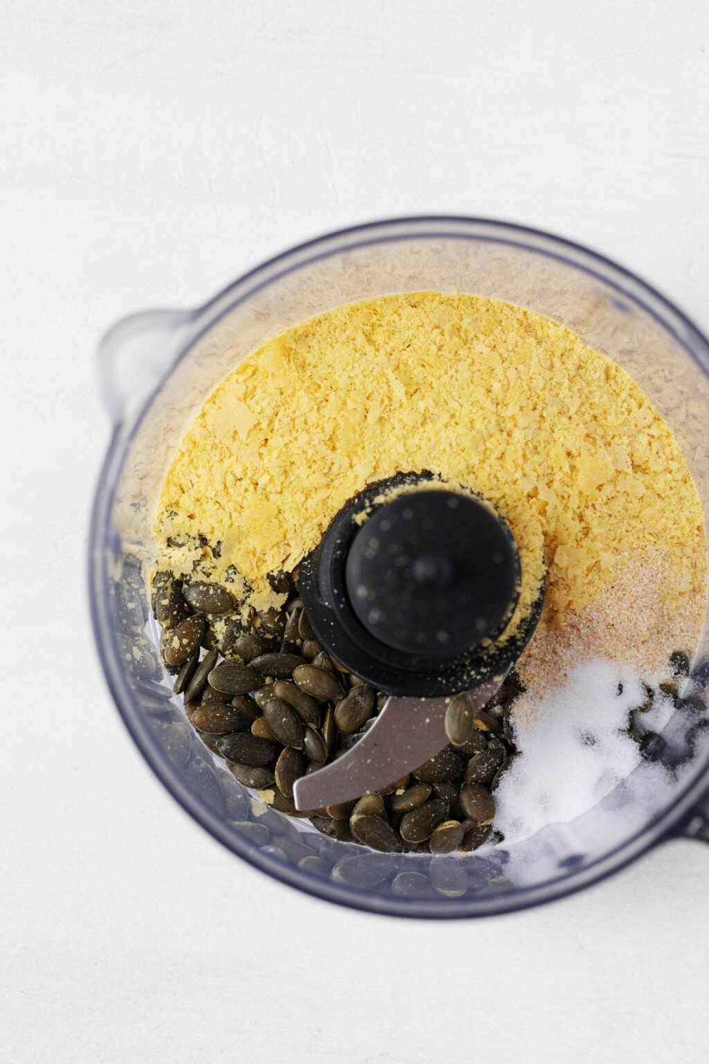 An overhead image of seeds and nutritional yeast in the clear container of a food processor.