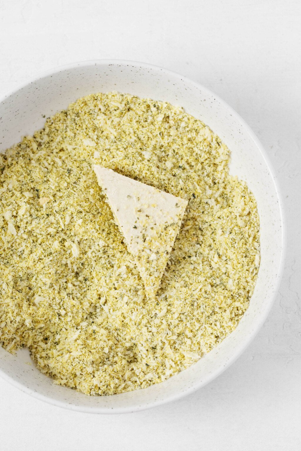 A triangle of tofu is being dipped into a finely ground mixture of nuts and nutritional yeast.
