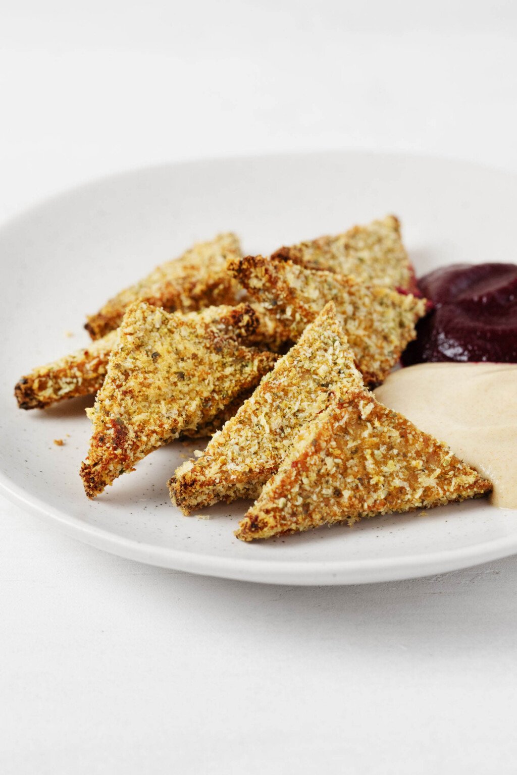 Triangles of pumpkin seed crusted tofu are arranged on a round, white plate. The tofu is served with one dark red and one cream-colored dipping sauce.