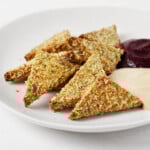 Triangles of pumpkin seed crusted tofu are arranged on a round, white plate. The tofu is served with one dark red and one cream-colored dipping sauce.