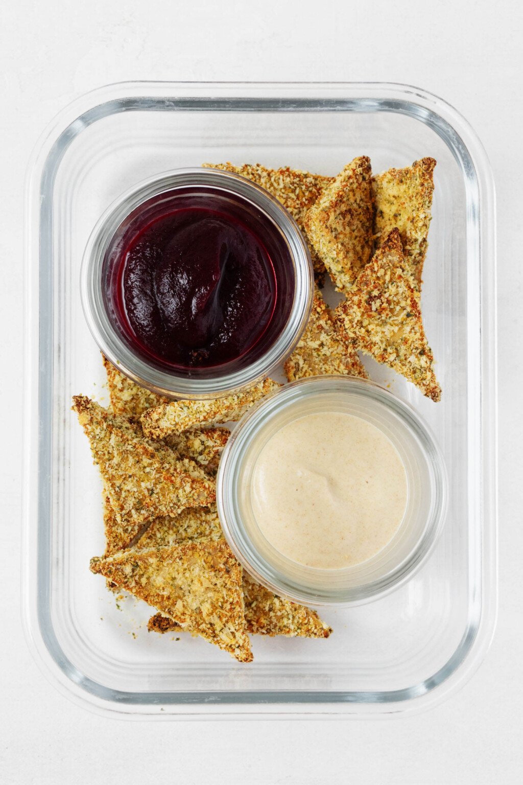 Triangles of pumpkin seed crusted tofu are arranged in a clear, rectangular storage container. The tofu is served with one dark red and one cream-colored dipping sauce.