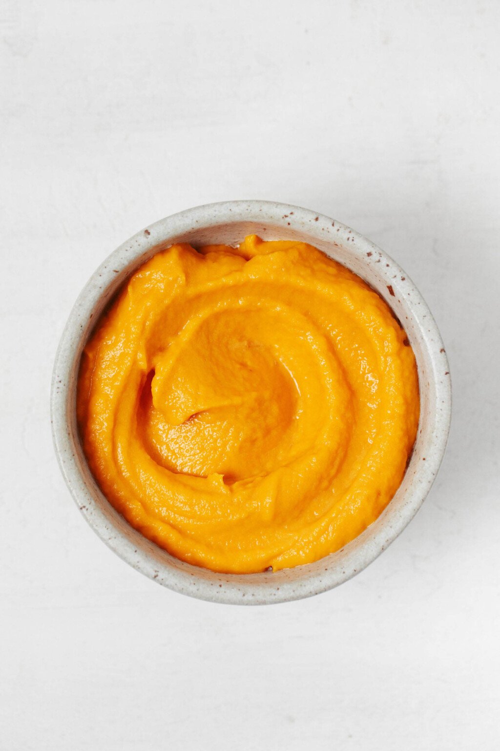 An overhead image of a small white bowl, filled with a creamy roasted red pepper spread and the corner of a cracker peeking out.