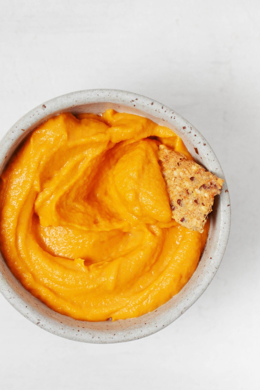 An overhead image of a small white bowl, filled with a creamy roasted red pepper spread and the corner of a cracker peeking out.
