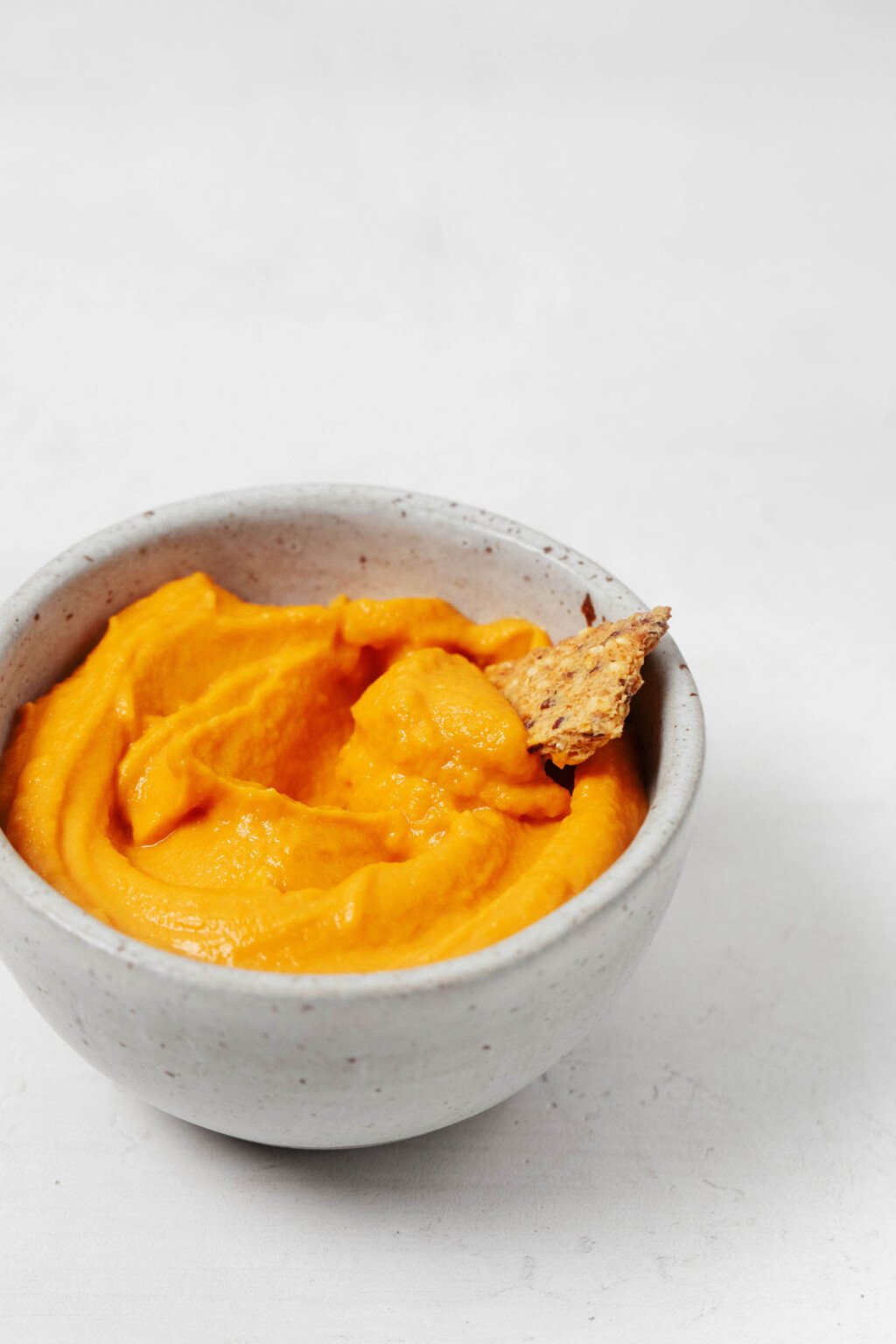 An overhead image of a small white bowl, filled with a creamy roasted red pepper spread and the corner of a cracker peeking out.