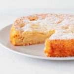An angled photograph of a vegan apple cake. One slice has been cut out, and the top has been dusted with powdered sugar.