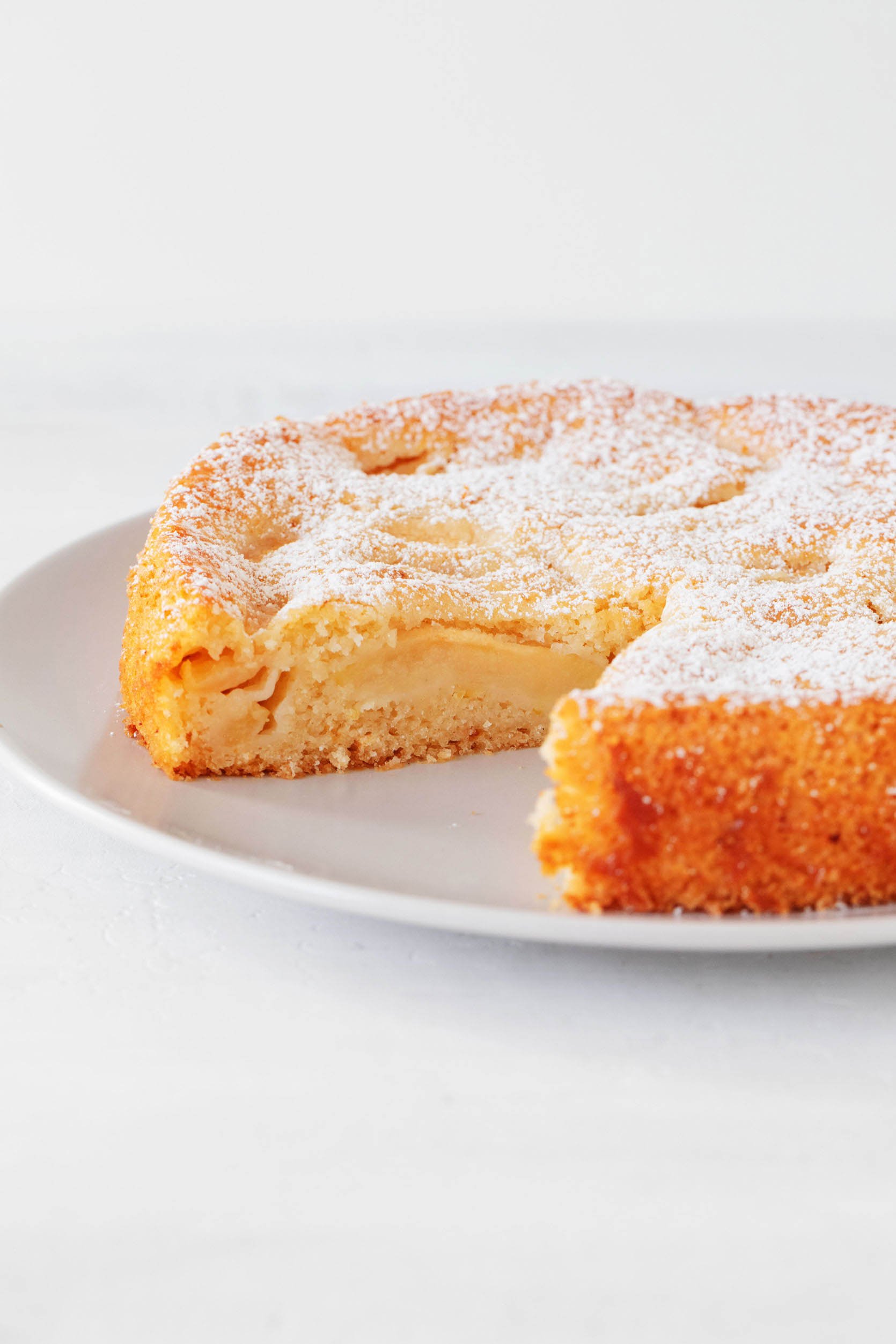 An angled photograph of a vegan apple cake. One slice has been cut out, and the top has been dusted with powdered sugar.