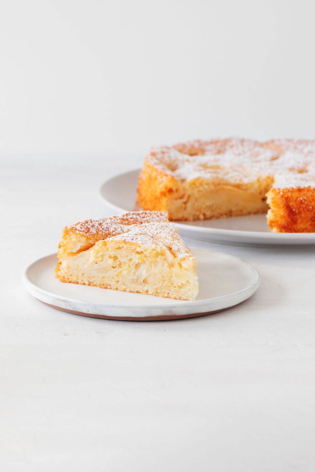 An Italian-inspired apple cake rests on a serving plate and is dusted with sugar. A single slice has been cut out of the cake and is being served on a smaller dessert plate.