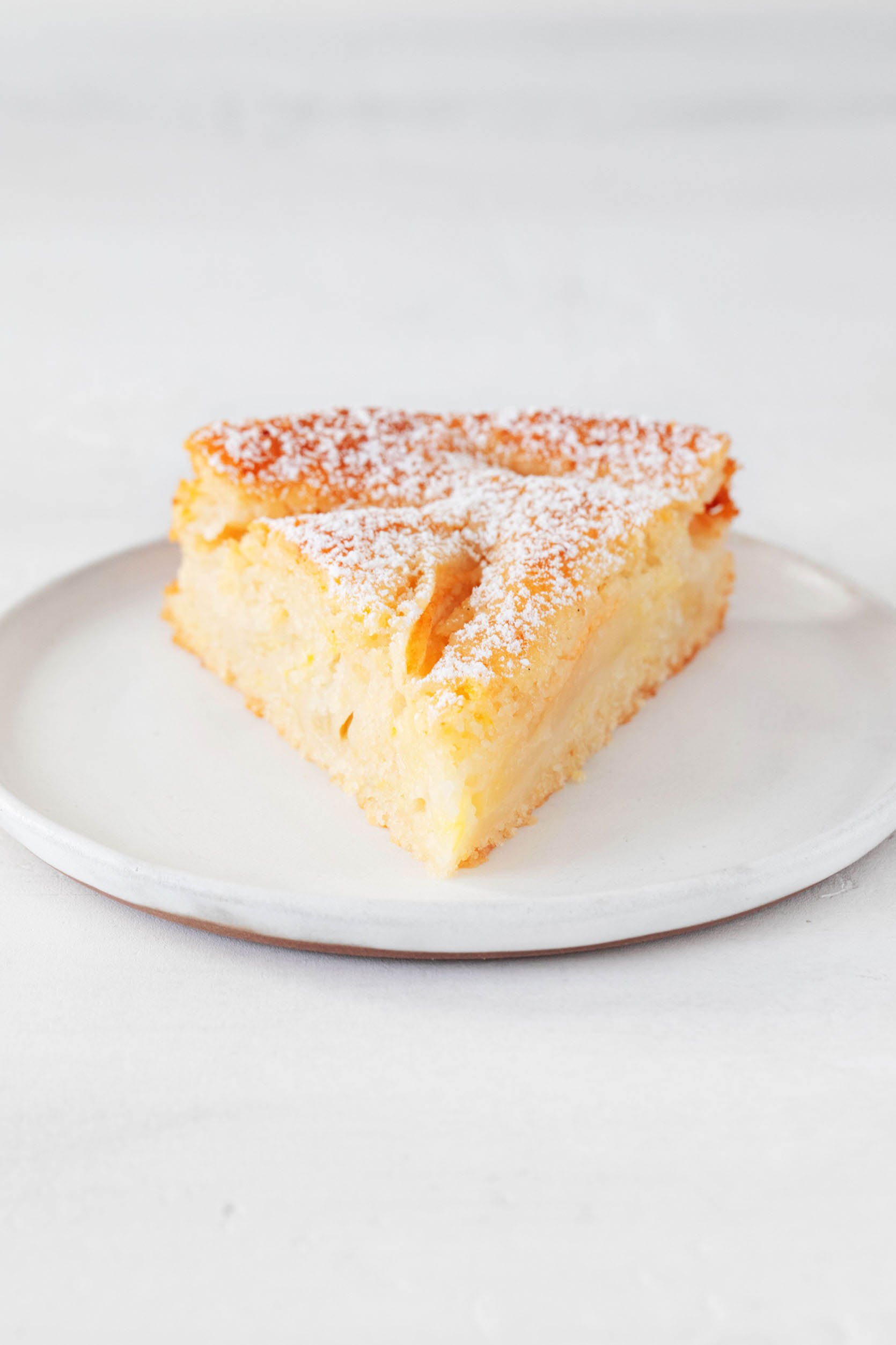 A single slice of apple cake, dusted with powdered sugar, rests on a round, small dessert plate. The plate is on a white surface.