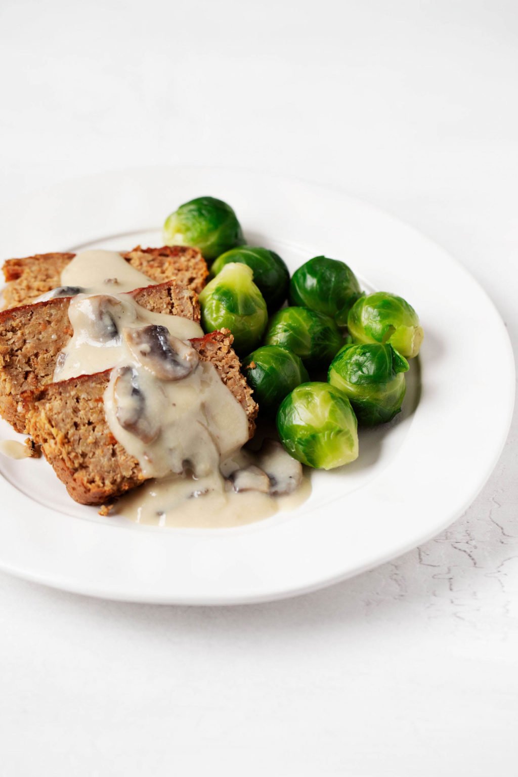 A vegan red lentil chickpea loaf has been topped with a creamy mushroom gravy. It's served on a white plate with Brussels sprouts.