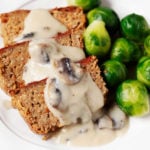 A round, white plate serves slices of red lentil chickpea loaf. Those slices are covered with mushroom gravy and served with bright green Brussels sprouts.