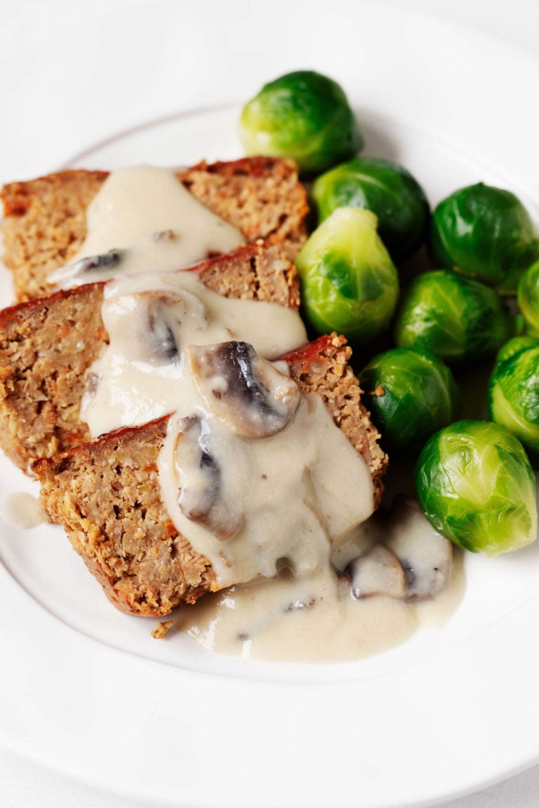 A round, white plate serves slices of red lentil chickpea loaf. Those slices are covered with mushroom gravy and served with bright green Brussels sprouts.