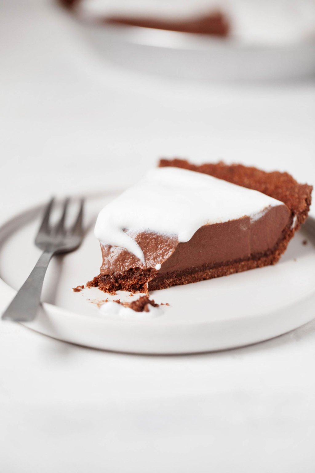 A slice of vegan chocolate mousse pie is topped with whipped cream. A fork rests alongside it on a small, white plate.