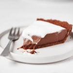 A slice of vegan chocolate mousse pie is topped with whipped cream. A fork rests alongside it on a small, white plate.