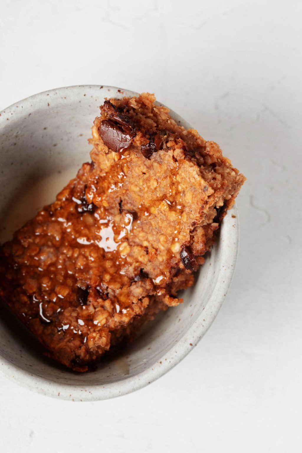 A small, ceramic bowl holds a slice of vegan pumpkin chocolate chip baked oatmeal, which is drizzled with maple syrup.