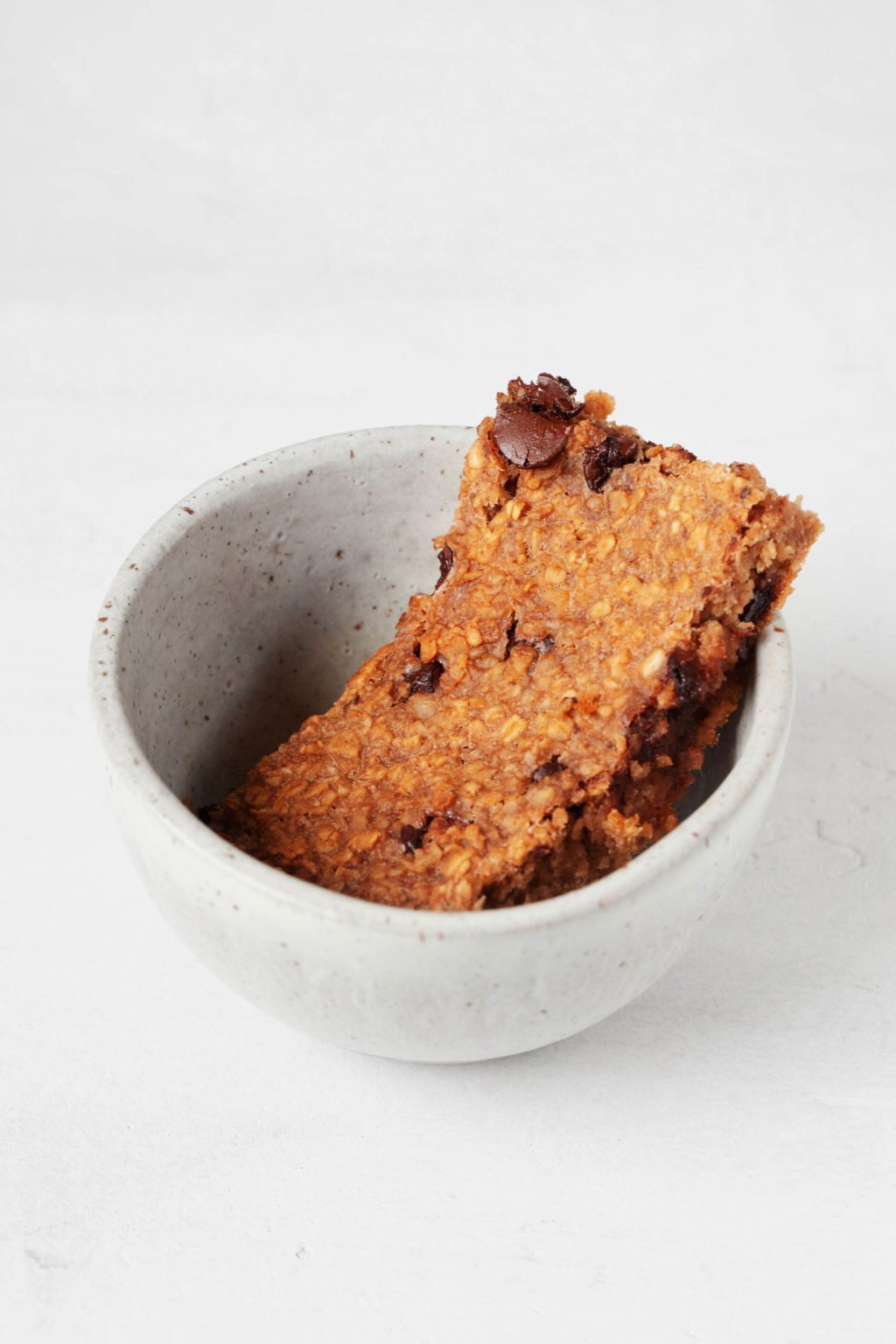 A small, white bowl is filled with a slice of a baked, whole grain breakfast that is dotted with chocolate chips.