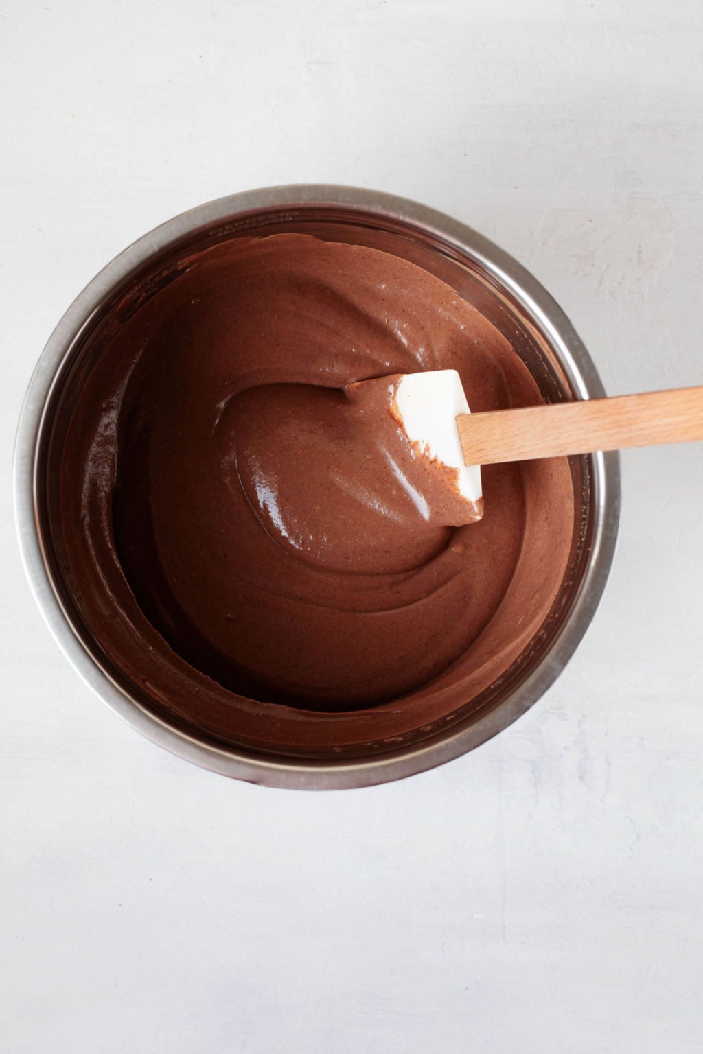 An overhead image of a silver bowl, which is filled with a creamy chocolate pudding mixture. A spatula stirs it. 