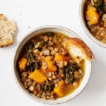 Two bowls of butternut kale lentil soup are laid on a white surface. A piece of toast rests nearby.