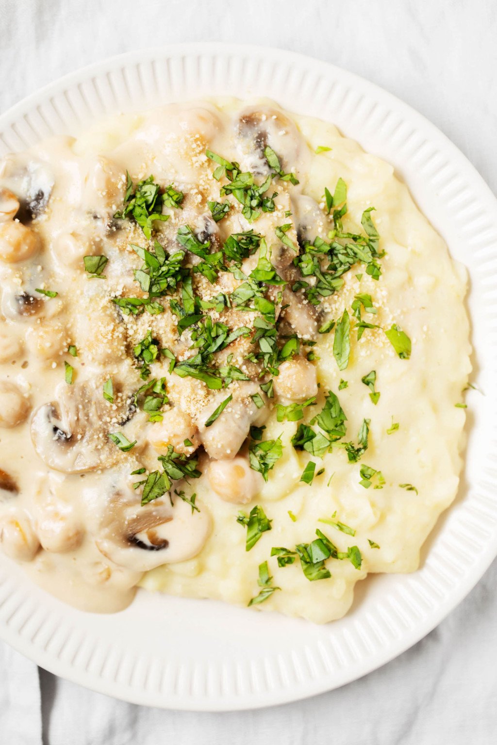 A white, fluted plate has been covered with a mixture of creamy chickpeas & mashed potatoes, then sprinkled with green herbs.