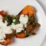 Two halves of sweet potatoes are topped with spiced lentils and a creamy tahini dressing. The potatoes rest on a round white plate.