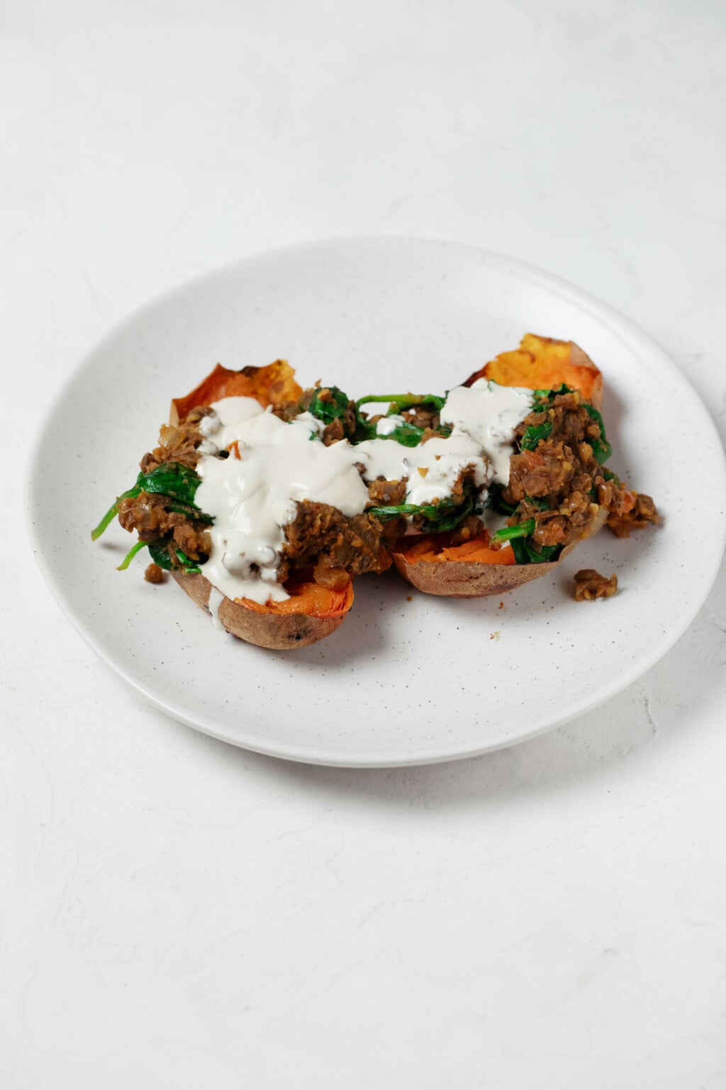 Two halves of sweet potatoes are topped with spiced lentils and a creamy tahini dressing. The potatoes rest on a round white plate.