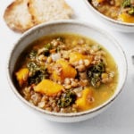 An angled photograph of two bowls of a vegan lentil, kale, and winter squash soup, which are served with toast points.