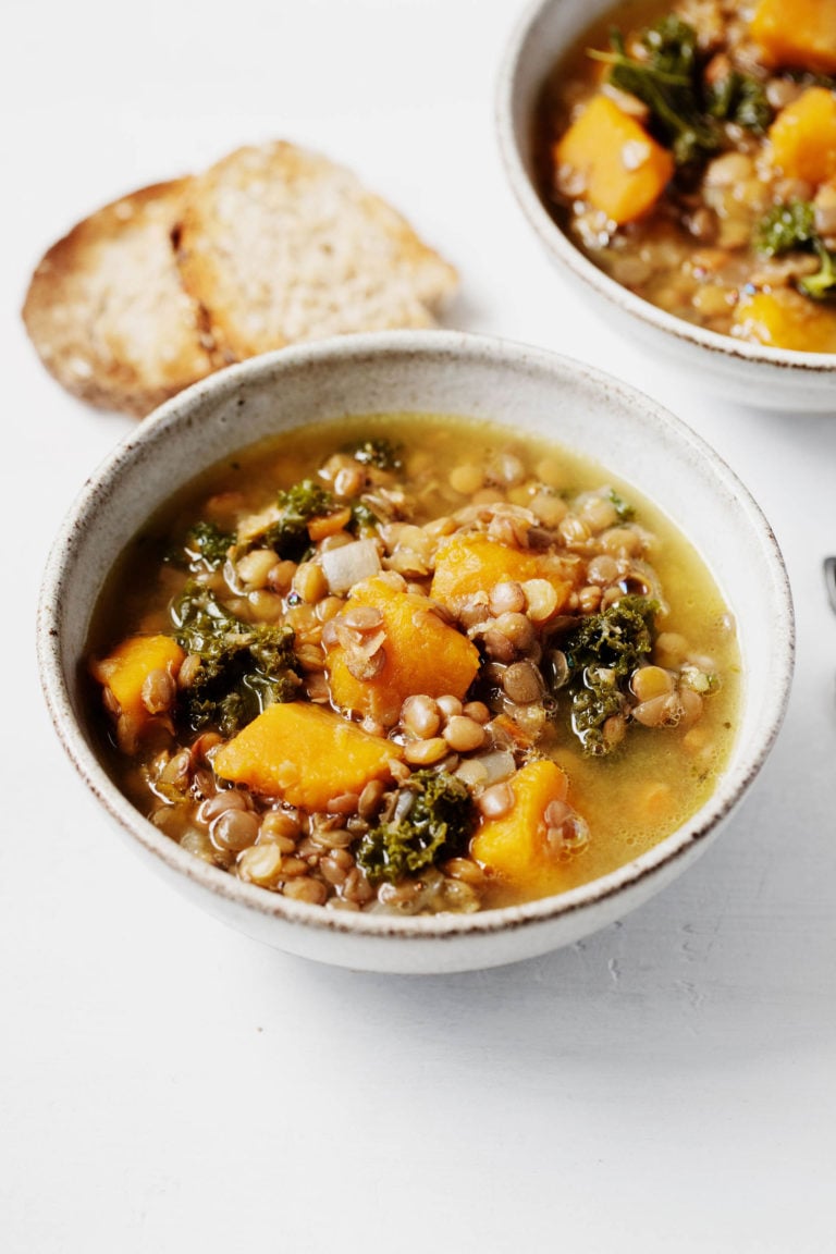 An angled photograph of two bowls of a vegan lentil, kale, and winter squash soup, which are served with toast points.