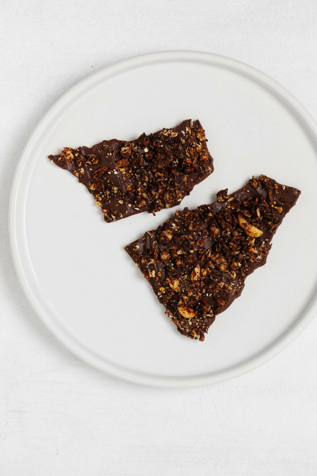Two pieces of vegan granola dark chocolate bark have been broken and placed on a small, round white plate.
