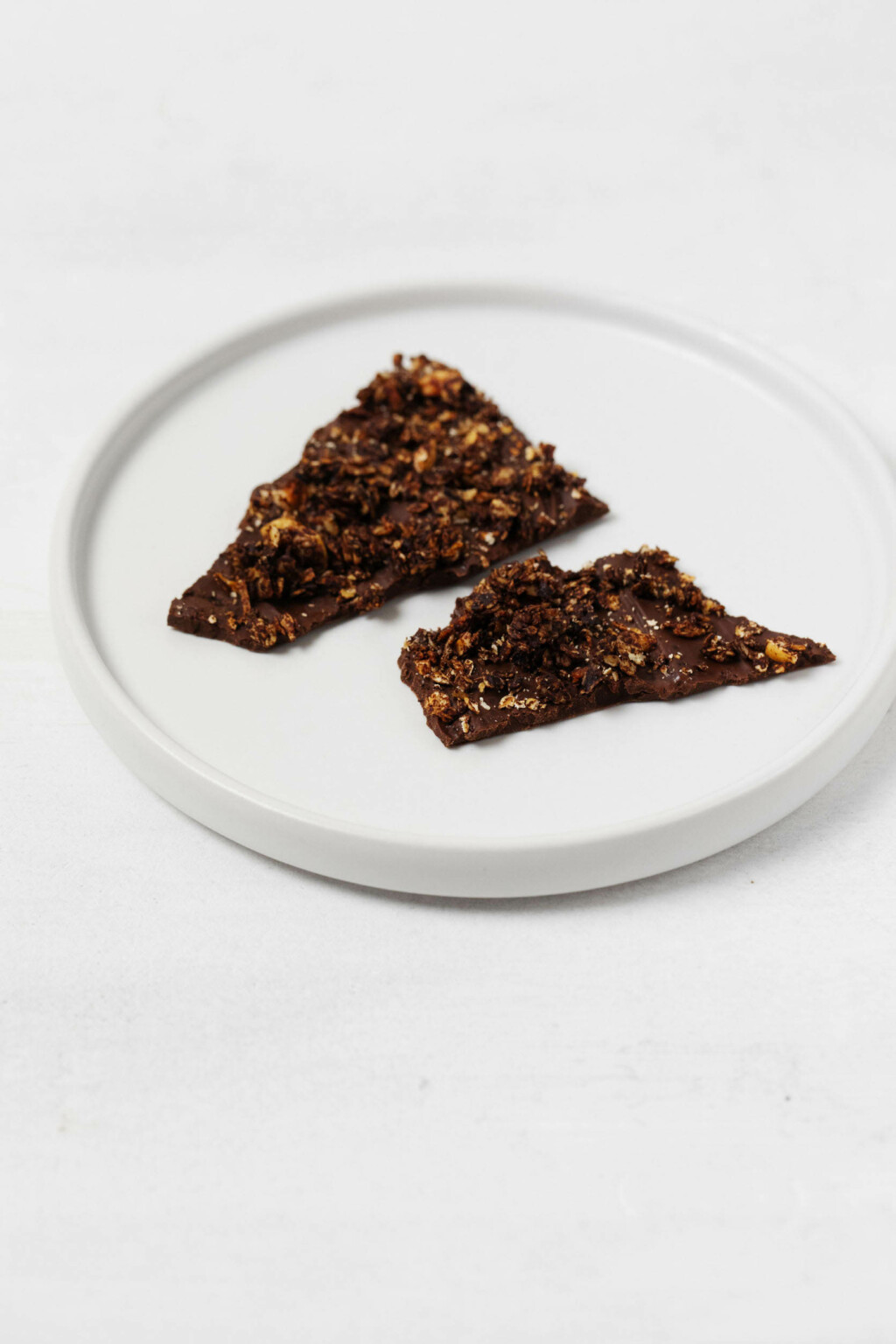 Two pieces of vegan granola dark chocolate bark have been broken and placed on a small, round white plate. The plate rests on a white surface.