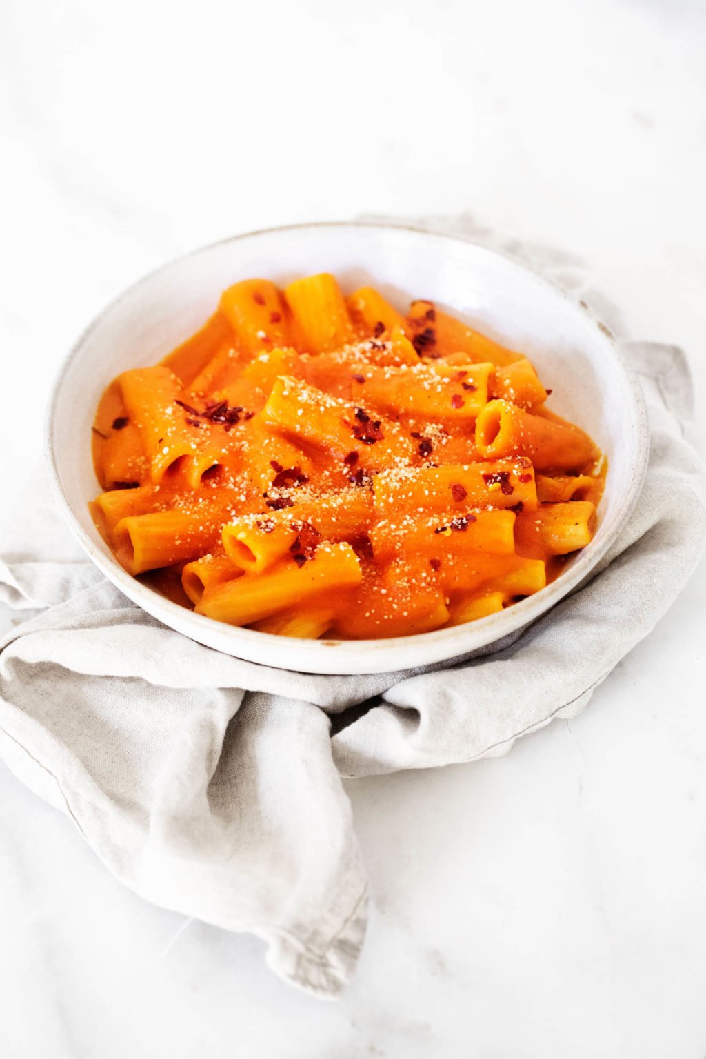 A white bowl, which is resting on a light gray linen against a white surface, has been filled with a creamy plant-based pasta alla vodka.