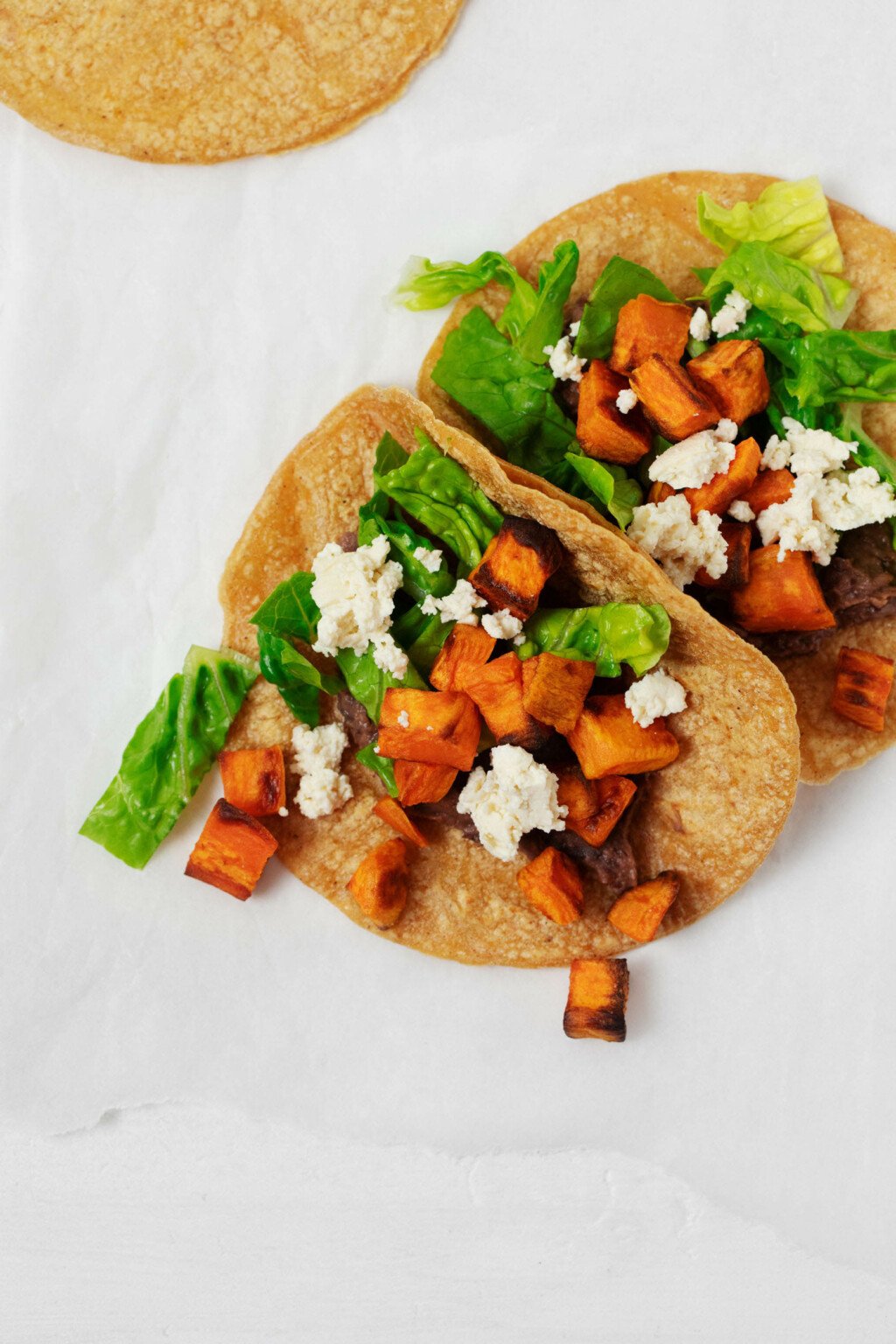 Vegan sweet potato tacos with black bean spread are plated on a white surface. They contain chopped lettuce.