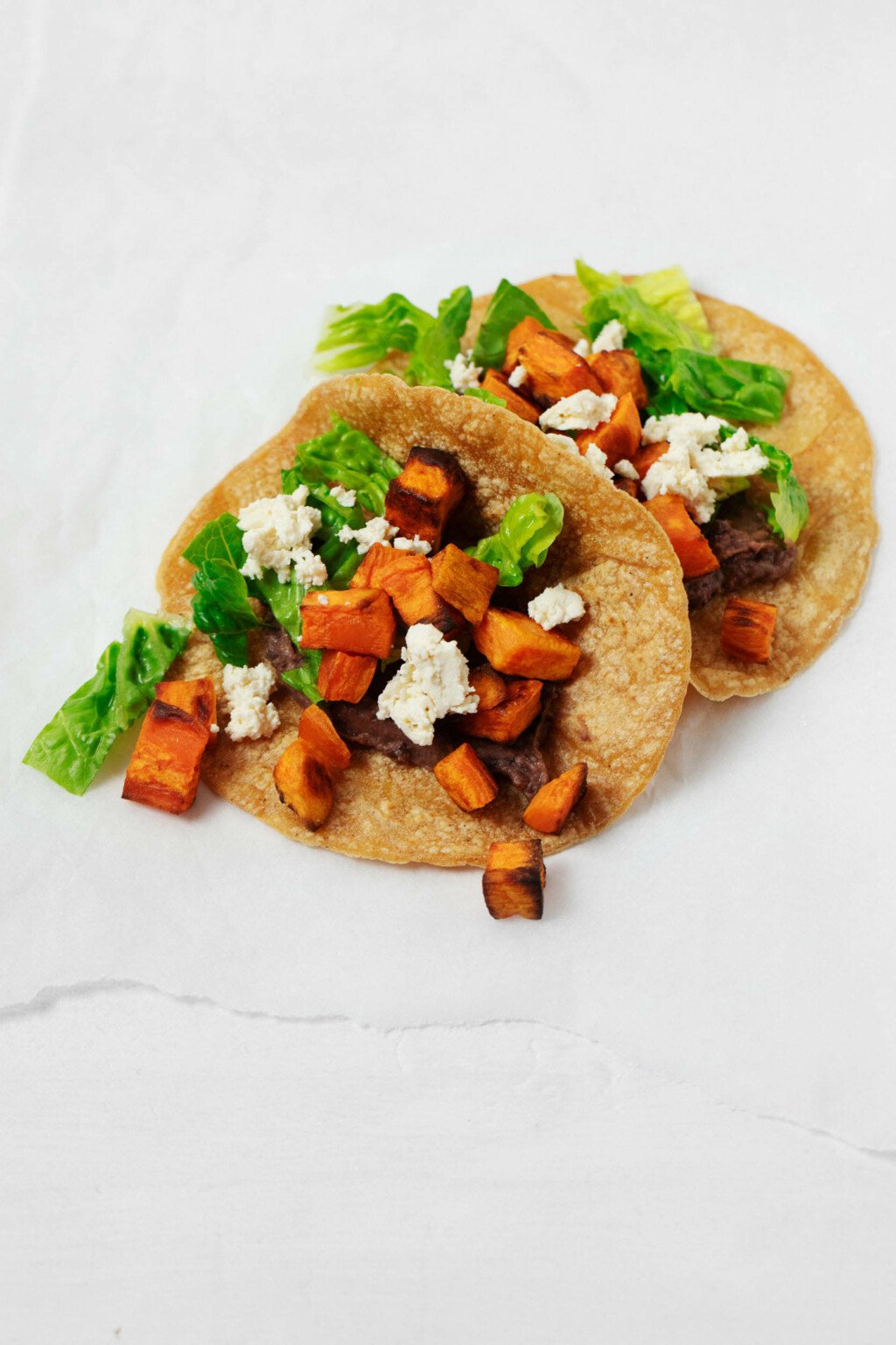 Two vegan sweet potato tacos with a black bean spread have been garnished with chopped lettuce and dairy free cheese.