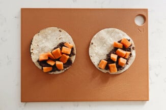 Tacos, made with a black bean spread and sweet potatoes, are being assembled on a rust-colored cutting board.