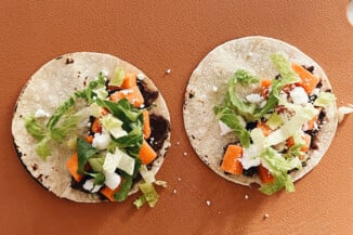 Sweet potato tacos with black bean spread have just been topped with green romaine lettuce and vegan cheese. They are resting on a cutting board.