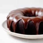 An angled photograph of a vegan chocolate bundt cake, which is placed on a serving platter and covered in a chocolate glaze.