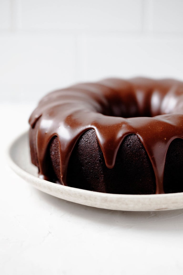 An angled photograph of a vegan chocolate bundt cake, which is placed on a serving platter and covered in a chocolate glaze.