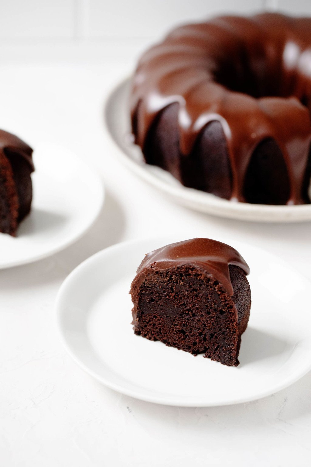 A slice of chocolate cake is on a small dessert plate in the foreground. The entire, glazed cake is sitting on a serving dish in the background.