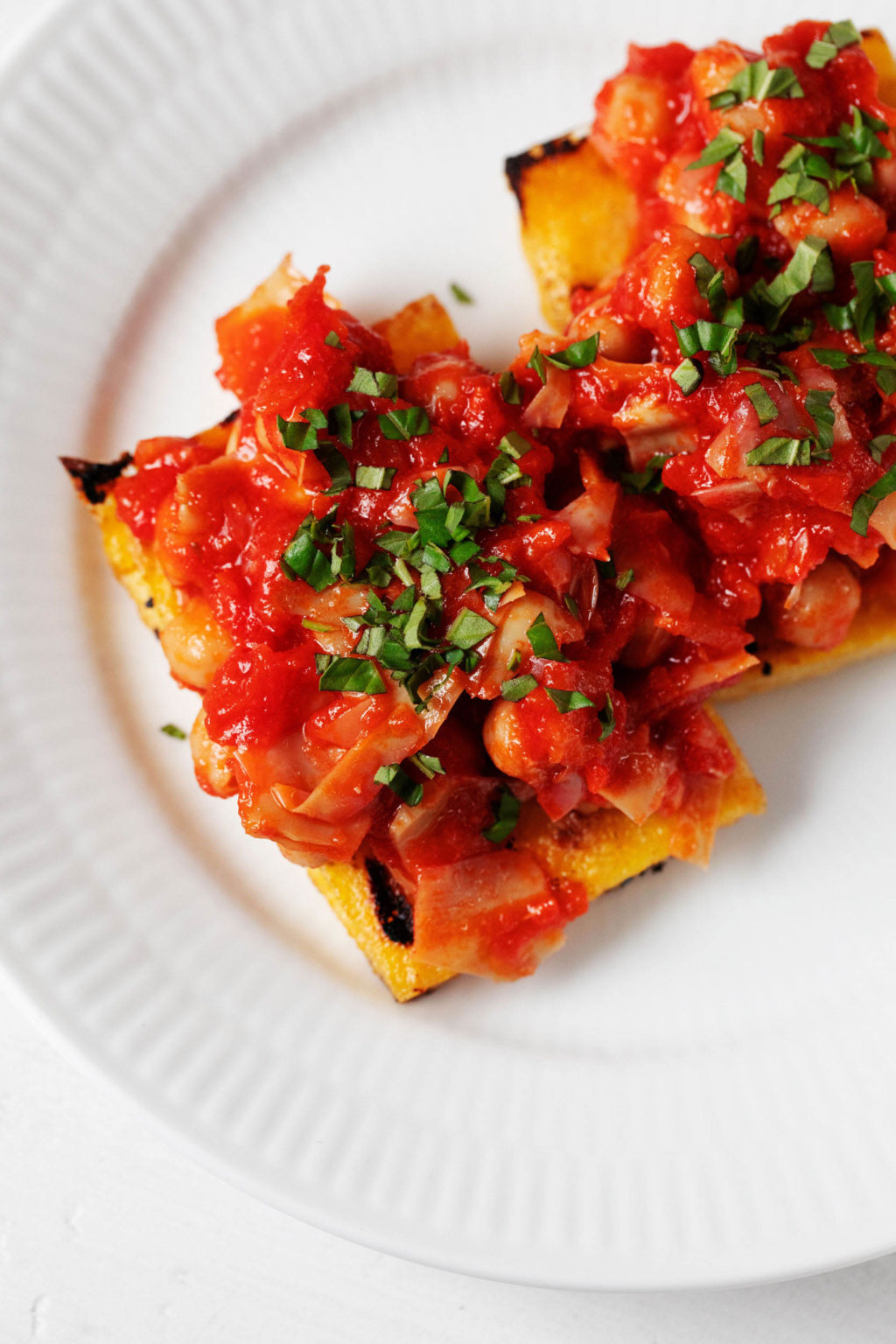 A saucy mixture of beans, vegetables, and tomatoes is piled on top of pieces of grilled polenta. They rest on a white plate on a white surface.